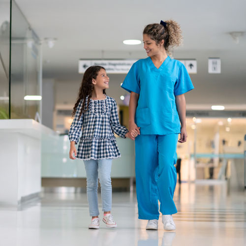 A woman physician and girl walking hand in hand and smiling down a hospital hallway