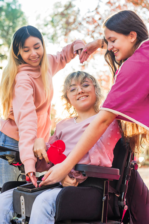 Two girls forming a heart shape with their arms around a girl in a wheelchair